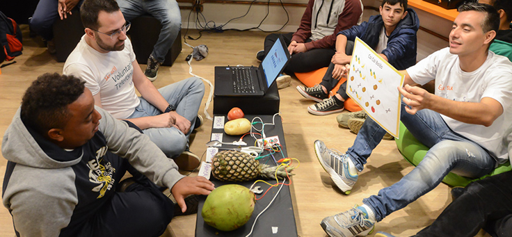 Voluntário está segurando placa que associa frutas conectadas a computador a sons em atividade que ensina programação a pessoas com deficiência intelectual.
