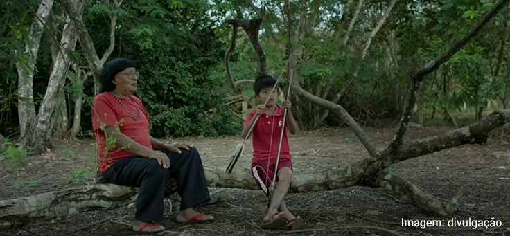 Imagem mostra cena do filme Ex-pajé, que retrata um pajé sentado em uma árvore e uma criança indígena manuseando algo que parece ser varetas