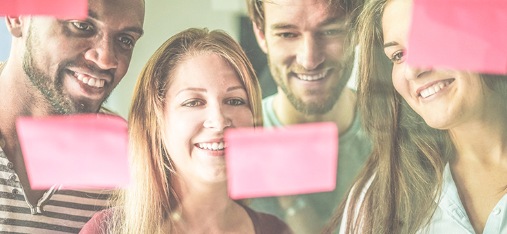 A imagem mostra quatro pessoas sorrindo enquanto olham para um quadro transparente em que se vê post-its colados.