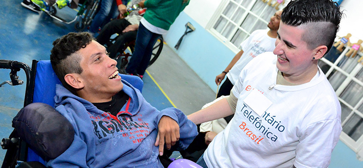 Imagem mostra voluntária conversando com pessoa com deficiência.
