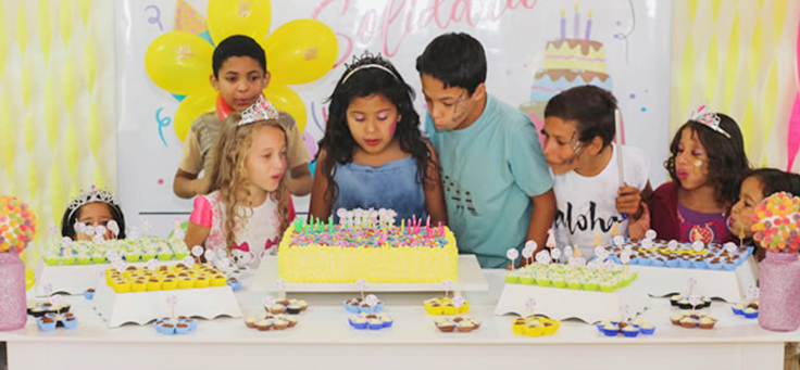 Crianças comemoram aniversário atrás da mesa, decorada com doces e bolo.