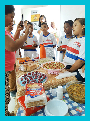 Imagem mostra alunos em volta de uma mesa com alimentos durante atividade de projeto sobre alimentação saudável.