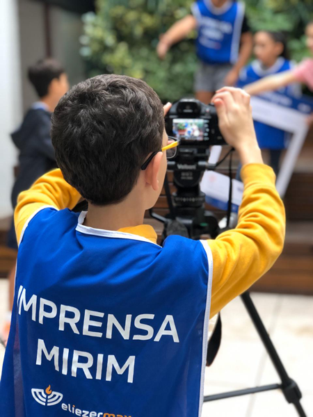 Imagem mostra aluno tirando fotos com uma câmera posicionada sobre um tripé. Ele usa um avental onde se lê “Imprensa Mirim”.