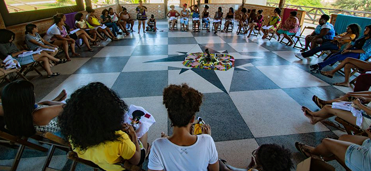 Cerca de 20 mulheres jovens e adultas estão sentadas em roda em um galpão com grandes janelas na Uneafro, iniciativa que contribui para a formação de jovens.