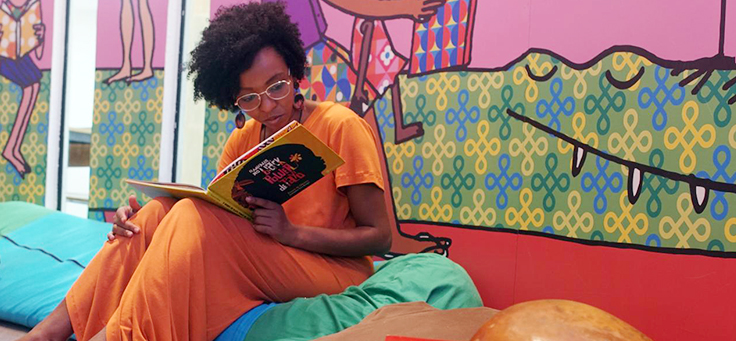 Imagem mostra mulher lendo livro. Ela está sentada sobre almofadas coloridas.
