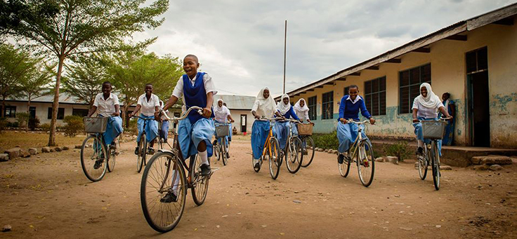 Imagem mostra um grupo de crianças uniformizadas andando de bicicleta em um chão de terra