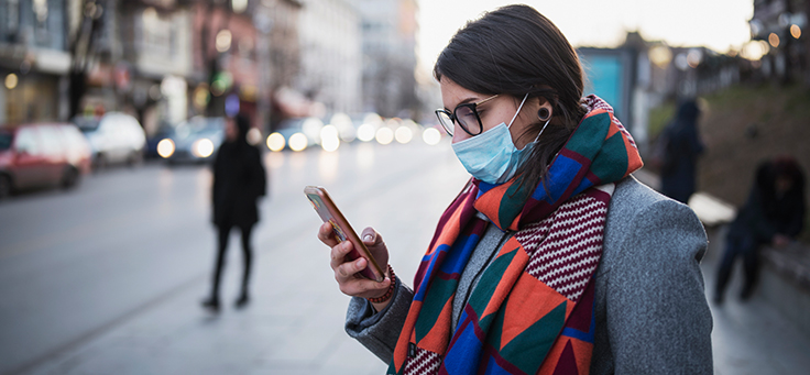 Imagem mostra uma mulher na rua usando uma máscara cirúrgica e vestindo casaco e cachecol. Ela está olhando para a tela de um celular que está em suas mãos.