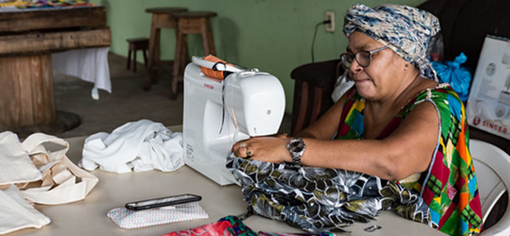 Mulher negra usando turbante e óculos está costurando peças em uma máquina para ilustrar negócios inseridos na economia solidária.