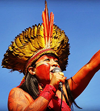 Célia Xakriabá é uma das lideranças indígenas que se destacam no Brasil. Ela está falando ao microfone e usando um cocar amarelo e laranja.