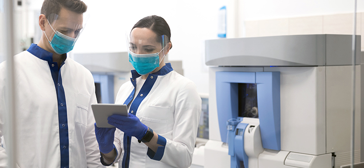 Imagem mostra um homem e uma mulher utilizando avental branco, luvas e máscara de proteção dentro de um laboratório