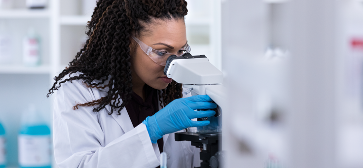 Imagem mostra uma mulher de jaleco e luvas olhando pela lente de um aparelho em um laboratório