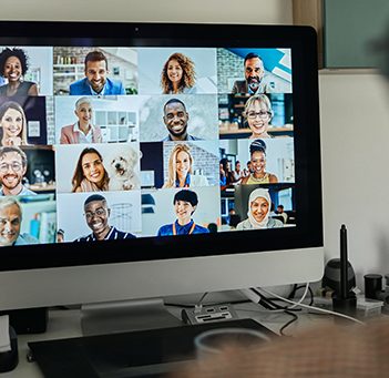 A imagem mostra uma pessoa de costas, sentada com fones de ouvido, olhando para a tela de um computador onde se vê o rosto de várias pessoas em uma videoconferência