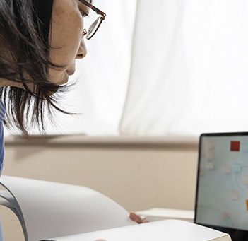 Mulher está olhando para tela de computador enquanto segura livro para ilustrar formação que une empreendedorismo, tecnologia e propósito. Ela usa óculos e tem os cabelos na altura do ombro.