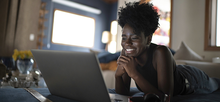 Imagem mostra uma jovem negra sorrindo com as mãos no queixo enquanto olha para a tela de um notebook