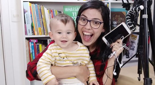 Lorena Carvalho, conhecida pelo canal Professora Coruja que foca em alfabetização e cultura digital, está segurando um bebê no colo e segurando um smartphone com a mão direita. Ao fundo há uma estante com livros.