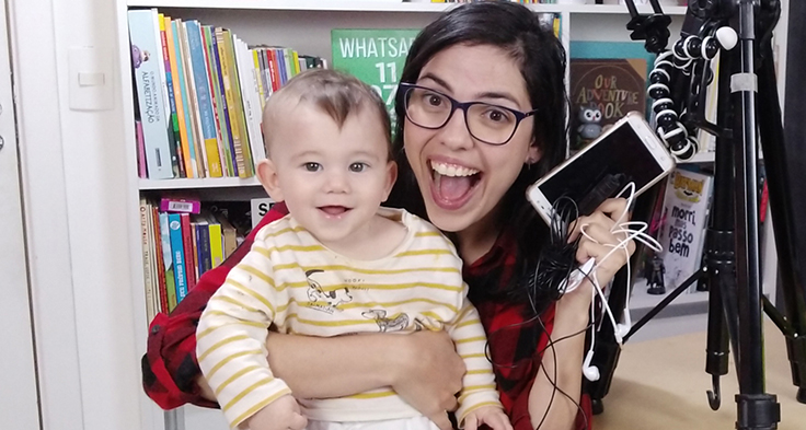 Lorena Carvalho, conhecida pelo canal Professora Coruja que foca em alfabetização e cultura digital, está segurando um bebê no colo e segurando um smartphone com a mão direita. Ao fundo há uma estante com livros.