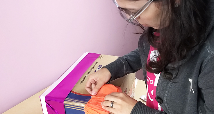 Lorena Carvalho, conhecida pelo canal Professora Coruja que foca em alfabetização e cultura digital, está de lado, colando um papel da cor laranja em uma caixa de papelão. Ela usa óculos e agasalho na cor cinza.