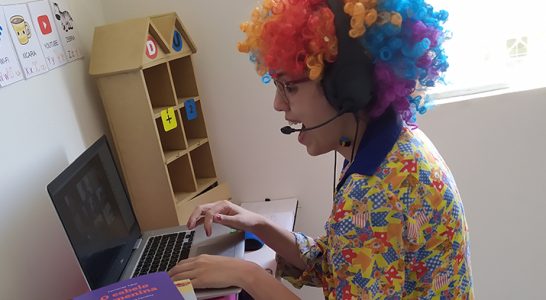 Lorena Carvalho, conhecida pelo canal Professora Coruja que foca em alfabetização e cultura digital, está digitando em um notebook enquanto veste uma peruca e uma roupa de palhaço.