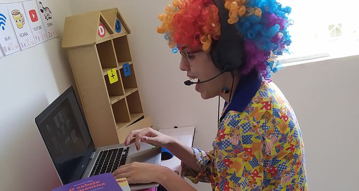 Lorena Carvalho, conhecida pelo canal Professora Coruja que foca em alfabetização e cultura digital, está digitando em um notebook enquanto veste uma peruca e uma roupa de palhaço.