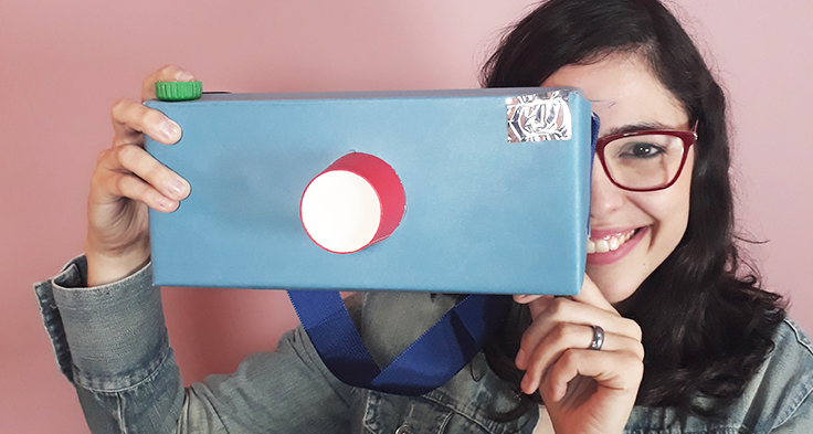 Lorena Carvalho, conhecida pelo canal Professora Coruja que foca em alfabetização e cultura digital, está segurando uma caixa de papelão encapada com papel colorido e simulando um modelo de câmera fotográfica.