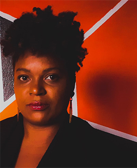 Foto de Magda Pereira de Souza, integrante do grupo Quem disse que eu não gosto de estudar?, que usou o pensamento computacional para criar um jogo relacionado à cultura afro-brasileira. Ela é uma mulher negra de cabelos enrolados, olha firme para a câmera e usa batom rosa e uma roupa preta.