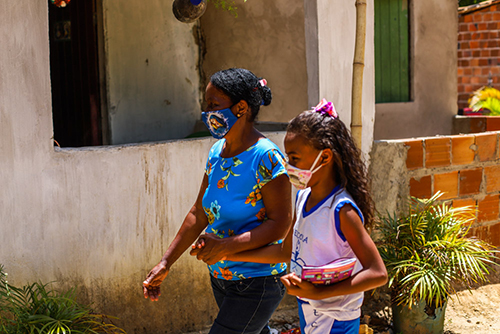 A imagem mostra uma senhora e uma criança de braços dados andando pela rua. Ambas estão com máscaras de proteção.