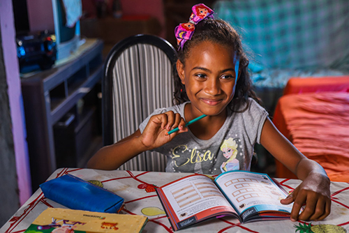 A imagem mostra uma menina sorrindo segurando um lápis e com um caderno a sua frente.