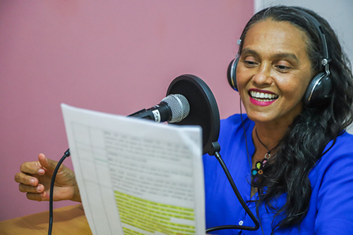 Imagem mostra uma mulher de camiseta azul e fones de ouvido lendo um papel enquanto fala ao microfone