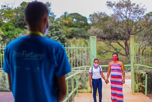 Imagem mostra uma mulher grávida de mãos dadas com uma menina de uniforme escolar e mochila, em frente a um portão aberto. Há um rapaz de costas, vestindo um camiseta azul, olhando para elas.