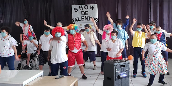 Beneficiários e colaboradores estão em pé dançando durante apresentação de show de talentos na Nova 4E durante a Semana dos Voluntários.