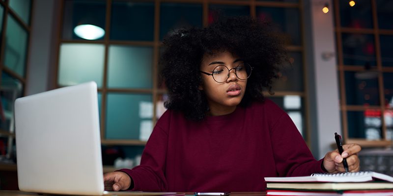 Imagem mostra mulher fazendo anotações em uma caderno em frente a um notebook.