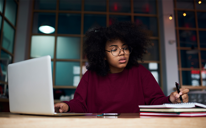 Imagem mostra mulher fazendo anotações em uma caderno em frente a um notebook.