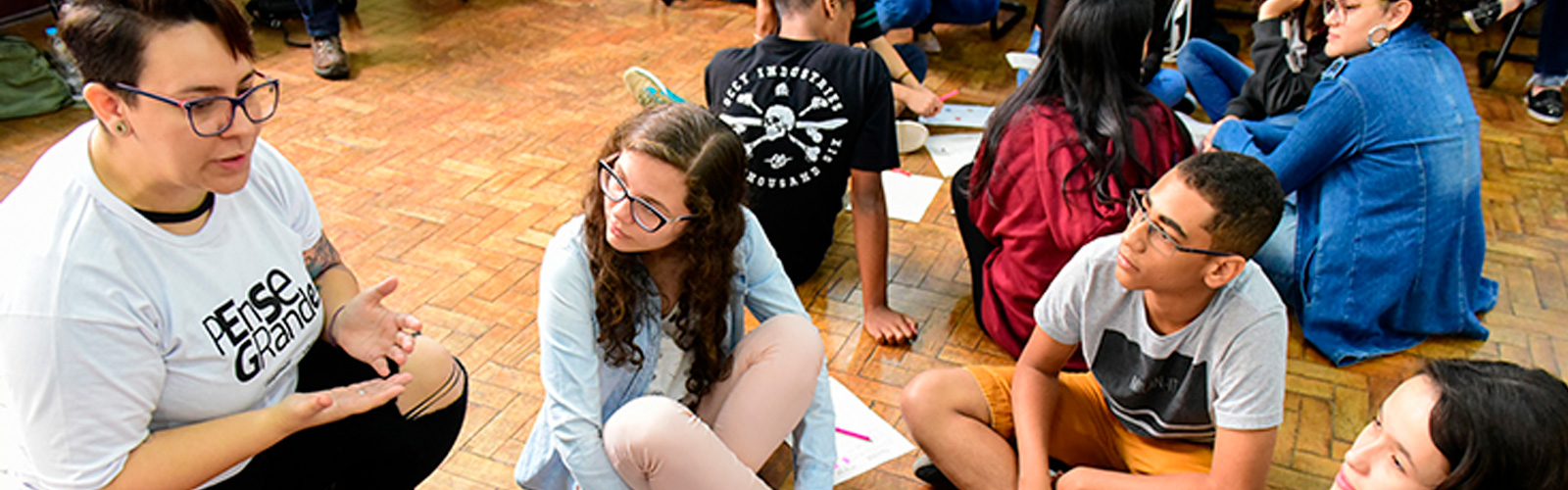 Jovens e formadores do programa Pense Grande estão sentados no chão em grupos, formando círculos.