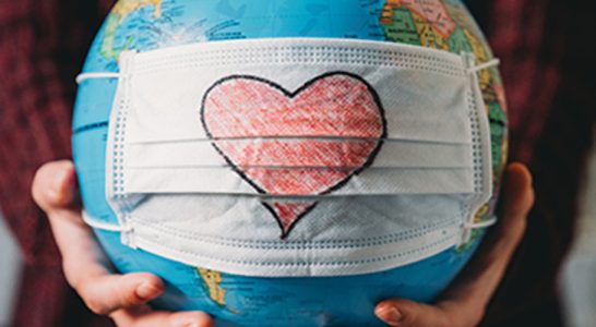 duas mãos seguram um globo terrestre que está com uma mascara de proteção contra a Covid-19 com um desenho de coração nela