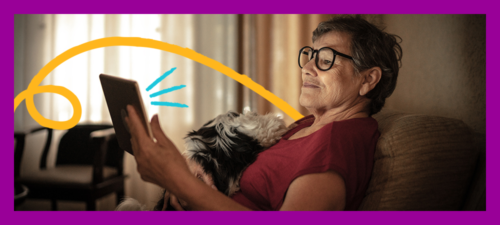 A imagem mostra uma senhora sentanda em um sofá com um cachorro no colo olhando para a tela de um tablet.