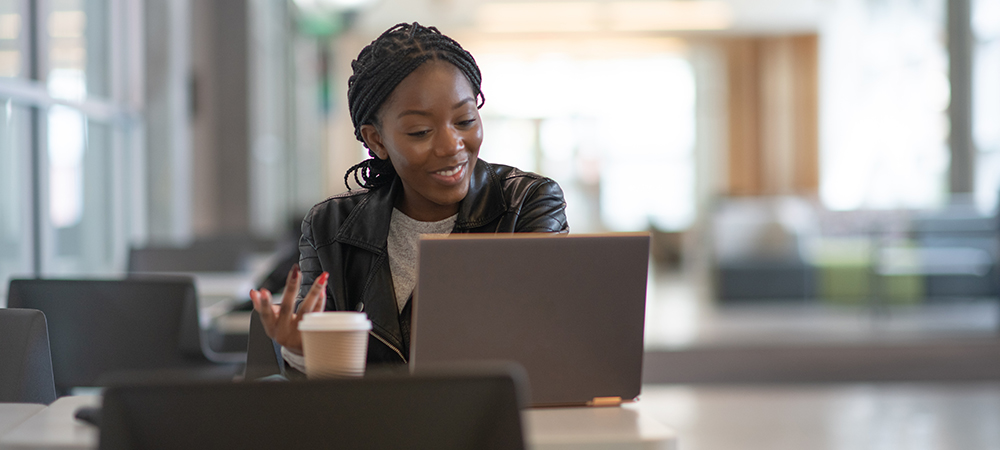 Imagem mostra uma jovem sentada atrás de uma mesa com um notebook a frente dela e um copo de café descartável ao lado