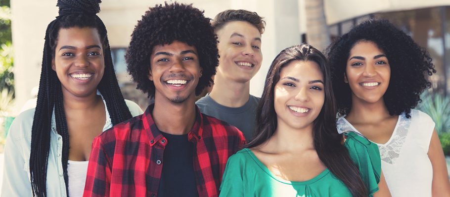 Imagem mostra um grupo de cinco jovens em pé, um ao lado do outro. Eles sorriem enquanto posam para a foto
