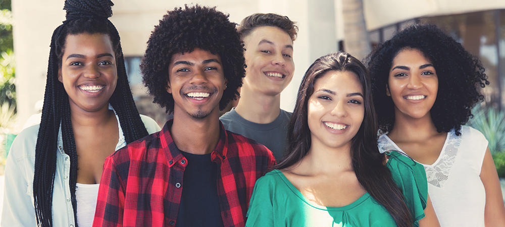 Imagem mostra um grupo de cinco jovens em pé, um ao lado do outro. Eles sorriem enquanto posam para a foto
