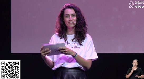 Mulher apresenta o evento feito a distância Cantata de Natal. Ela tem os cabelos ondulados na altura do ombro e usa a camiseta com os dizeres: eu sou voluntário Telefônica Vivo.