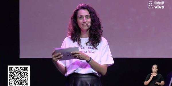 Mulher apresenta o evento feito a distância Cantata de Natal. Ela tem os cabelos ondulados na altura do ombro e usa a camiseta com os dizeres: eu sou voluntário Telefônica Vivo.