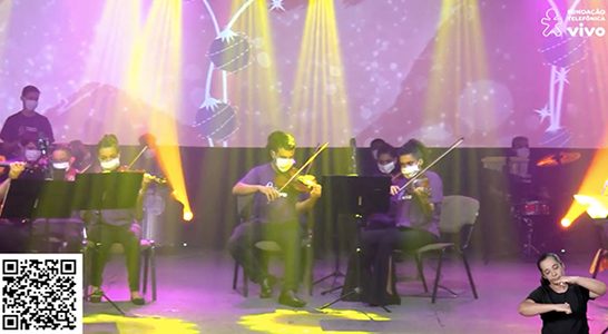 Imagem mostra jovens tocando violino durante o espetáculo Cantata de Natal. Todos estão em cima de um palco, usando máscara de proteção facial e camiseta roxa com logo da Vivo.