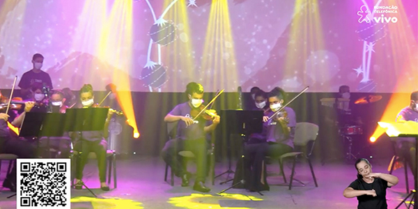 Imagem mostra jovens tocando violino durante o espetáculo Cantata de Natal. Todos estão em cima de um palco, usando máscara de proteção facial e camiseta roxa com logo da Vivo.