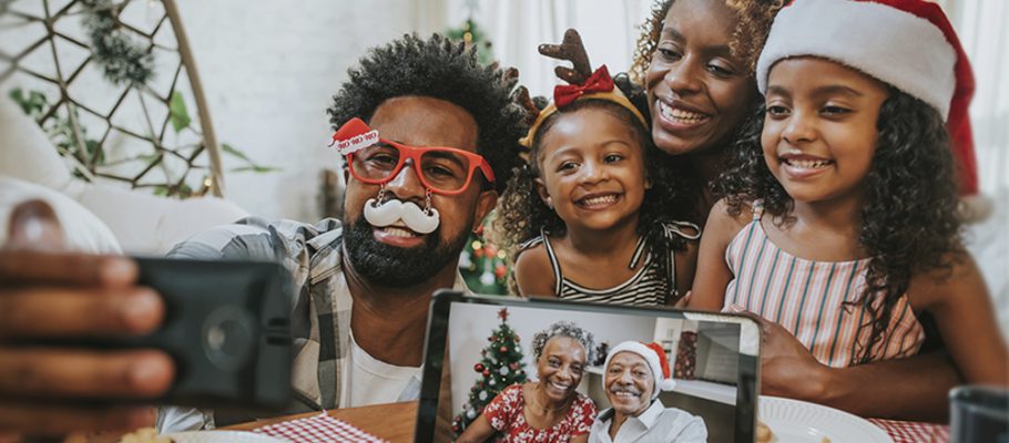 Imagem mostra pai, mãe e filhas fazendo chamada com avós durante o Natal. Os avós aparecem em uma tela de tablet e todos os usam adereços natalinos, como gorro de Papai Noel.