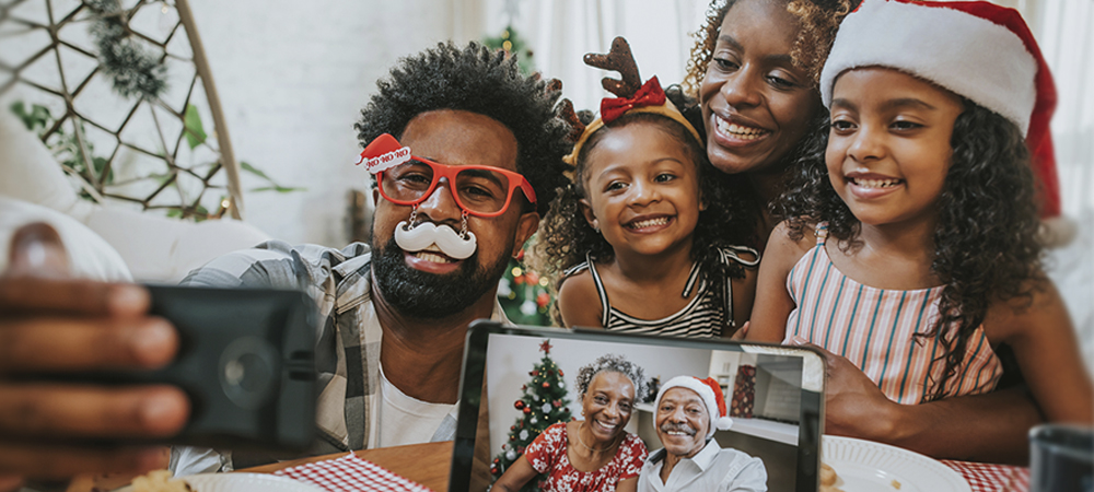 Imagem mostra pai, mãe e filhas fazendo chamada com avós durante o Natal. Os avós aparecem em uma tela de tablet e todos os usam adereços natalinos, como gorro de Papai Noel.