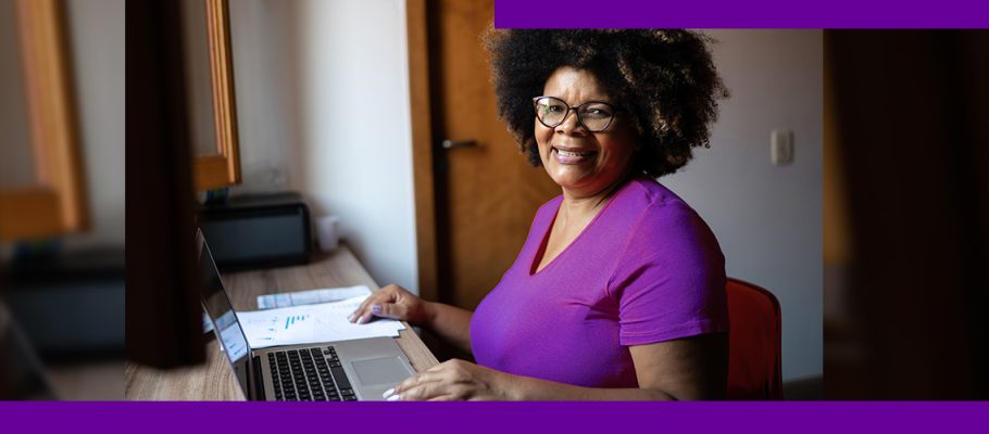 Imagem mostra uma senhora negra sentada em frente a uma mesa com as mãos sobre um notebook. Ela usa uma camiseta púrpura, óculos de grau e está sorrindo.