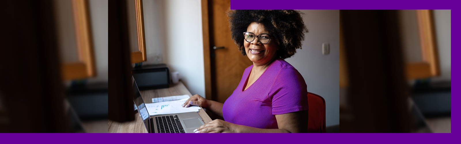 Imagem mostra uma senhora negra sentada em frente a uma mesa com as mãos sobre um notebook. Ela usa uma camiseta púrpura, óculos de grau e está sorrindo.