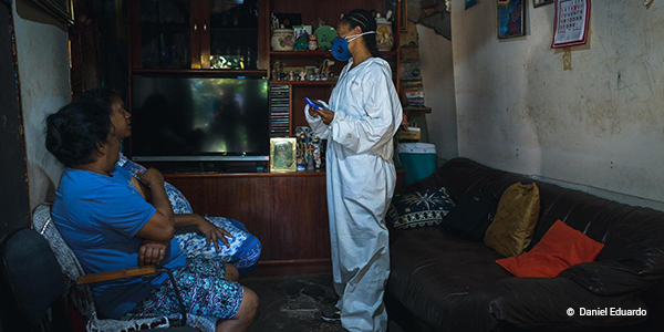 A imagem mostra uma voluntária de máscara e roupa de proteção branca dentro da casa de duas mulheres em Paraisópolis. Elas estão sentadas olhando para a voluntária que está de pé.