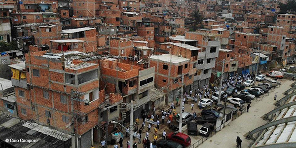 A imagem, vista de cima, mostra um grupo de pessoas andando pelas ruas de Paraisópolis. É possível ver várias casas e construções de tijolos.