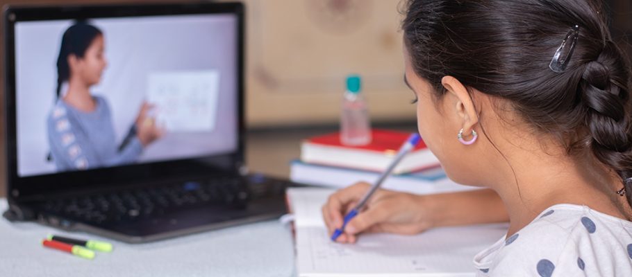 Imagem mostra uma menina sentanda de costas escrevendo em um caderno. Sobre a mesa, há alguns livros e um notebook com a imagem na tela de uma mulher segurando um papel branco.