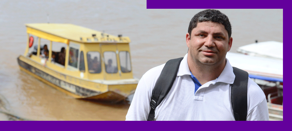 O Prof. Gilson da Silva, que atuou em escola atendida dentro do projeto Aula Digital, está sorrindo e, ao fundo, barcos se movimentam por rio que corta a cidade de Manaus (AM).